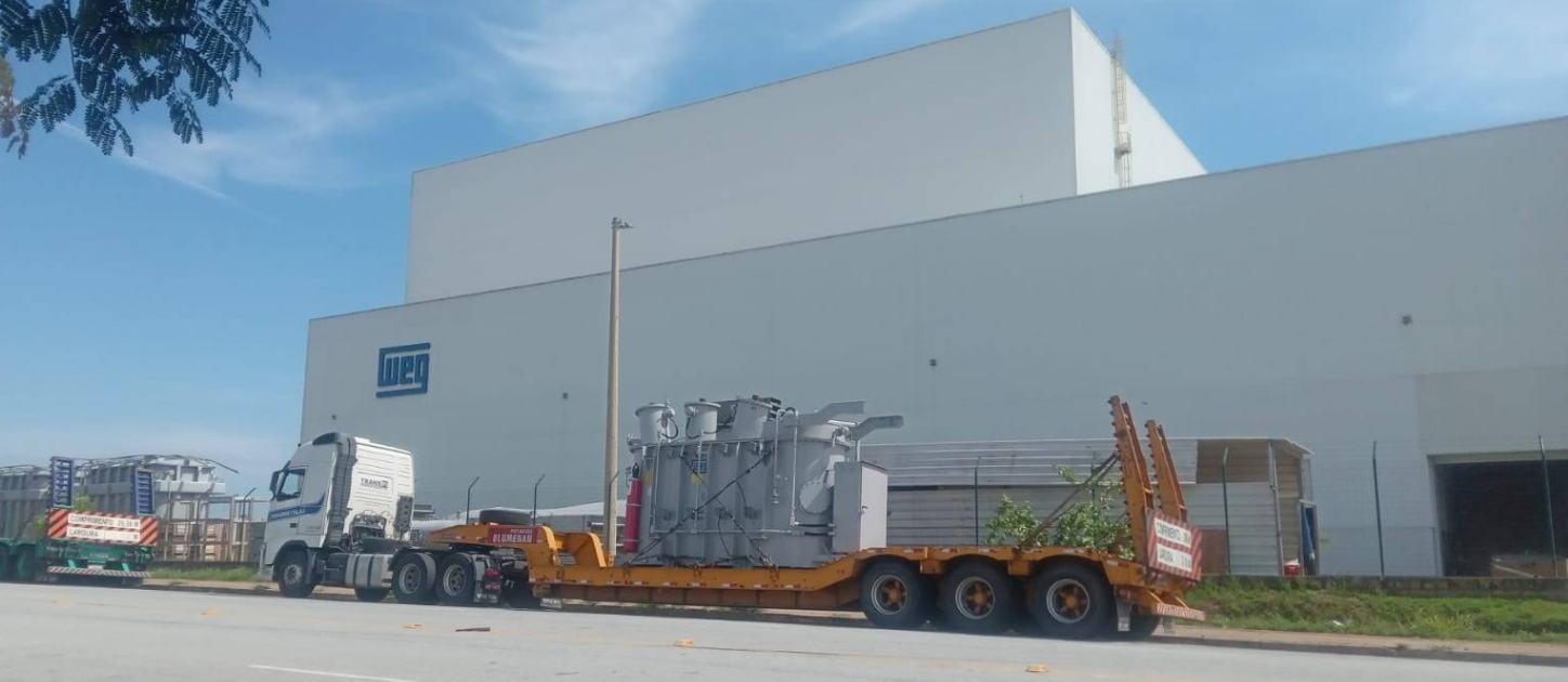 AO FUNDO O GALPÃO INDUSTRIAL DA WEG FÁBRICA DE TRANSFORMADORES COM UM CAMINHÃO EMBARCANDO UM TRANSFORMADOR DE GRANDE PORTE À FRENTE - PROJETO INDUSTRIAL DA RENATO MELO ARQUITETURA
